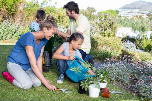 A family working together in a well-maintained garden in Pimlico, with lush green grass, flowering plants in flower beds, and garden tools scattered around. The foreground features a woman kneeling on the grass, planting or tending to a small shrub, with a child nearby holding a watering can filled with greenery. Behind them, a man and a young girl are seated on the lawn, engaging in a gardening activity. The background includes a mix of flowering bushes, shrubs, and small trees, creating a vibrant and inviting outdoor space. The garden appears to be in good condition, with neatly bordered flower beds and a lush, healthy lawn, reflecting professional gardening and landscaping services typical of Pimlico. Natural daylight enhances the scene, suggesting a clear, pleasant day suitable for outdoor maintenance. This image exemplifies family-friendly gardening activities that a local company like Landscapers Pimlico might support through their landscape enhancement services, ensuring vibrant, functional garden spaces in the area.