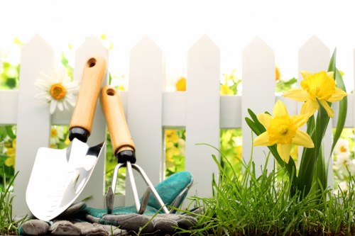 This image depicts a section of a well-maintained garden with a white picket fence in the background, highlighting the outdoor environment typical of residential landscaping in Pimlico. In the foreground, bright yellow daffodils and white daisies grow amidst lush green grass, which appears dense and healthy with varied shades of green, indicating good lawn care practices. The garden features a narrow flower bed border along the fence, filled with vibrant plants, while the soil area adjacent to the grass is dark and well-turned, suggesting recent planting or soil maintenance. Resting on the ground are gardening tools including a small hand trowel with a white blade, a hand rake, and a pair of gardening gloves, all positioned amidst the greenery, indicating ongoing or recent gardening activity. The scene is illuminated by natural daylight, with a soft lighting that enhances the natural tones of the plants and the wooden tools, creating a peaceful and inviting outdoor space suitable for garden landscaping and maintenance services provided by Landscapers Pimlico, aimed at enhancing residential gardens in the local area around postcode SW1.