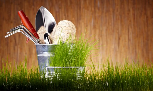 The image features a close-up view of gardening tools and supplies arranged outdoors on a lush green lawn. In the foreground, there are two small metal buckets placed on the grass; one contains a bundle of fresh, bright green grass and three gardening tools with red and silver handles, including a trowel, a hand rake, and a small fork. The second bucket holds a ball of natural twine, suitable for tying and supporting plants. Behind these buckets, the garden surface transitions into a wooden background that appears to be part of a garden shed or fence, with warm tones and a smooth texture. The overall scene appears to be set on a bright, partly cloudy day, ideal for outdoor gardening activities. This outdoor arrangement showcases typical gardening equipment and natural elements that landscapers Pimlico might incorporate into their garden design and maintenance services, emphasizing a well-kept and inviting outdoor space in a residential setting near Pimlico, London, with natural lighting highlighting the garden’s inviting atmosphere.