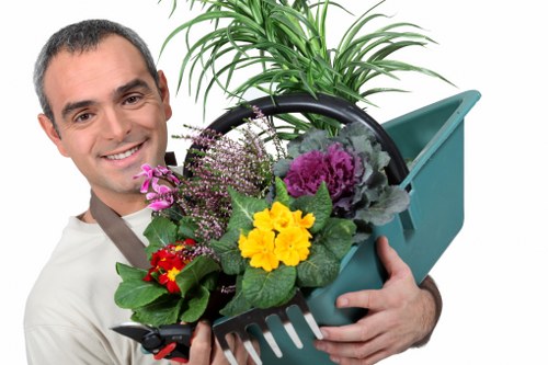 The image depicts a smiling man in a gardening apron holding a green plastic garden basket filled with a variety of colourful flowering plants, including purple, yellow, and red blooms, along with lush green foliage. In the background, there is a well-maintained garden with a neatly trimmed lawn, bordered by a low hedge, and a tall, leafy plant. The outdoor space appears to be part of a residential garden in Pimlico, with clear weather and natural daylight illuminating the scene. The scene emphasizes outdoor gardening and planting activities typical of professional garden maintenance or landscaping services such as those offered by Landscapers Pimlico, supporting the context of lawn and garden care within a small urban garden setting.