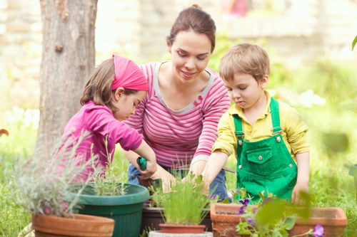 A woman with dark hair pulled back, wearing a pink striped long-sleeve shirt, is engaged in gardening outdoors in a backyard, accompanied by two young children. One child, wearing a purple jacket and pink headband, is using a gardening tool to tend to a potted plant, while the other child, in a yellow shirt with green overalls, observes nearby. They are surrounded by various potted plants with lush green foliage and vibrant purple and yellow flowers. The garden features a large tree trunk in the background, with a wooden fence and blurred greenery creating a natural, inviting environment. The scene captures a sunny day, with soft natural light illuminating the garden space, highlighting the different textures of the soil, plant leaves, and flower petals. This outdoor area is well-maintained, with a mix of garden beds and pots that reflect typical features of a landscaped residential garden in Pimlico, supporting gardening and outdoor maintenance services provided by Landscapers Pimlico.