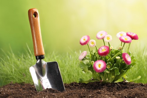 A close-up view of a gardening scene in a residential outdoor space, featuring a trowel with a wooden handle partially embedded in dark, freshly turned soil. To the right, there is a vibrant cluster of pink and white flowers with rounded petals and green foliage, planted in a neat flower bed border. The background showcases a well-maintained grassy lawn with a blurred green and yellow gradient, suggesting a sunny day. The overall setting indicates an outdoor garden or backyard in Pimlico, suitable for lawn care, planting, and gardening services by Landscapers Pimlico, with natural lighting emphasizing the lush greenery and bright floral elements.